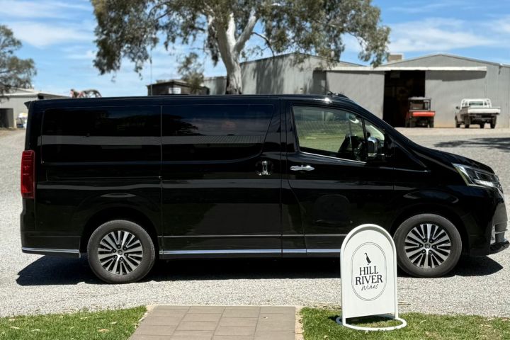 Black van parked on gravel near a 'Hill River Wines' sign, with shed and tree in the background.