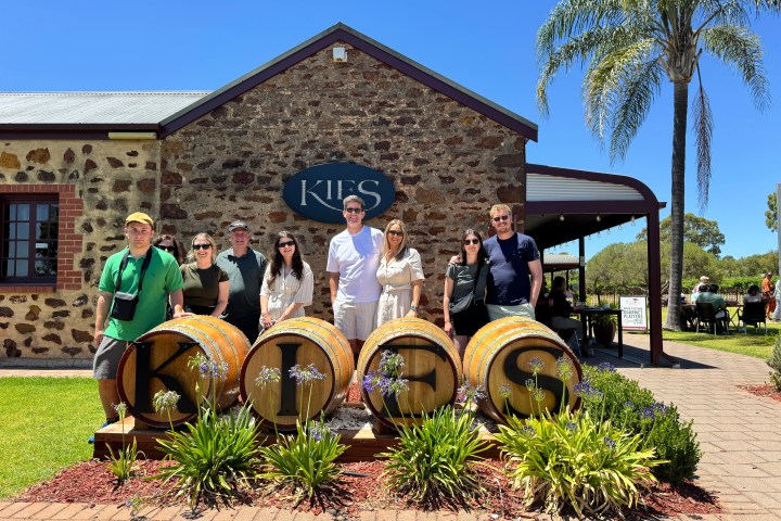 Group of people standing in front of Kies winery, with barrels and a palm tree on a sunny day.