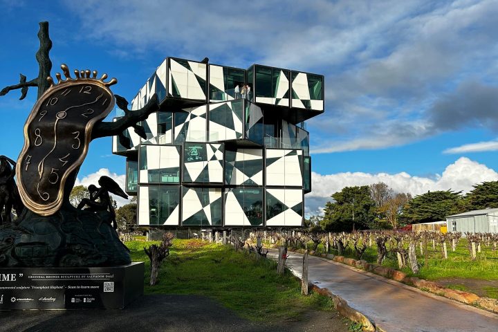 Modern cube building with a surreal sculpture in front, under a cloudy blue sky.