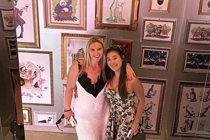 Two women in dresses standing by a wall with colorful framed art and mirrors.
