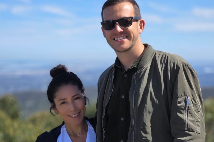 Two people posing outdoors on a sunny day with greenery and blue sky in the background.