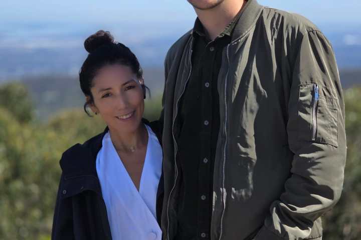 Two people posing outdoors on a sunny day with greenery and blue sky in the background.