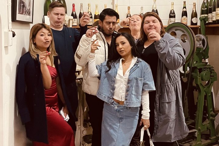 Group of six people posing indoors with a portrait and wine bottles on shelves.