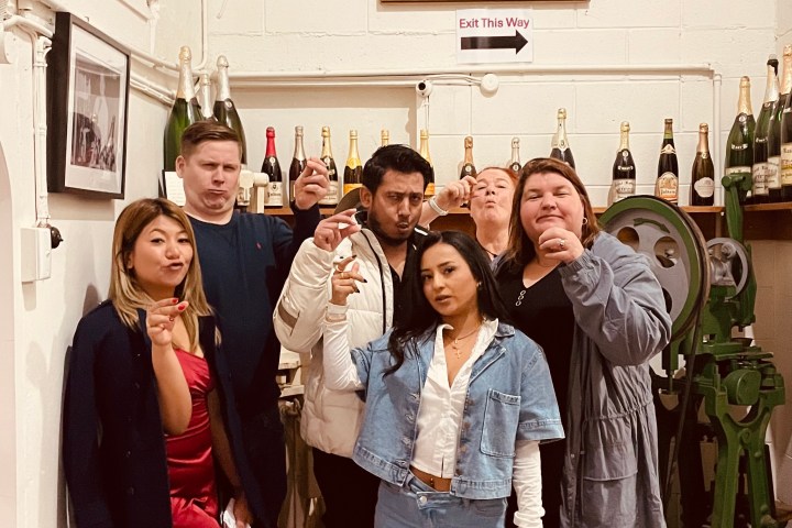 Group of six people posing indoors with a portrait and wine bottles on shelves.