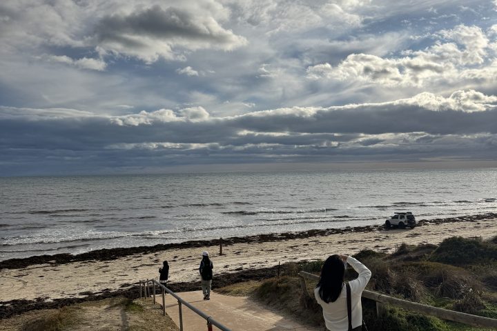 Two people walking towards a beach under a cloudy sky, with a car visible near the shoreline.