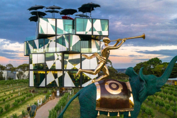 Modern building with geometric patterns, surrounded by vineyards and elephant statue, sky with scattered clouds.