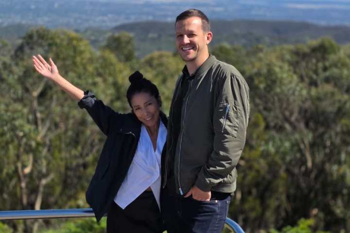 Two people smiling at a scenic overlook with trees and a distant city view.