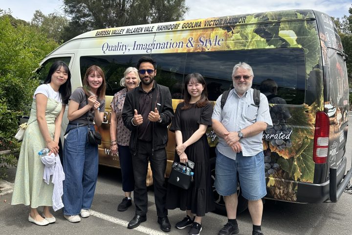 Six people stand in front of a tour van with vineyard branding, trees in the background.
