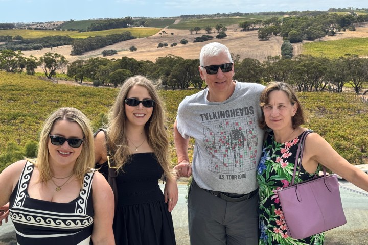 Four people wearing sunglasses pose in front of a vineyard landscape on a sunny day.