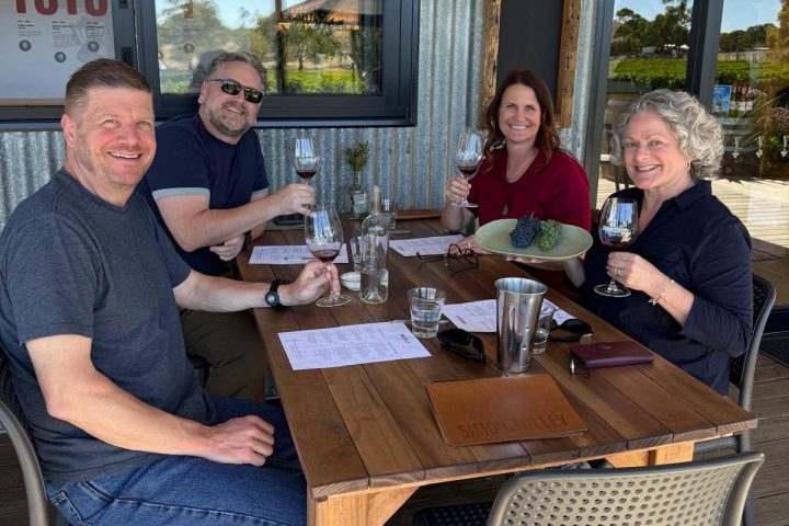 Four people sitting at an outdoor table, smiling and holding wine glasses.