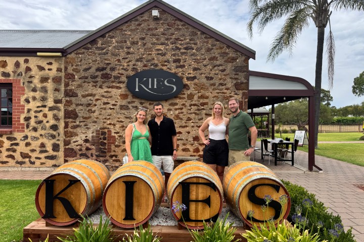 Four people stand in front of a stone building with wine barrels spelling 'KIES'.