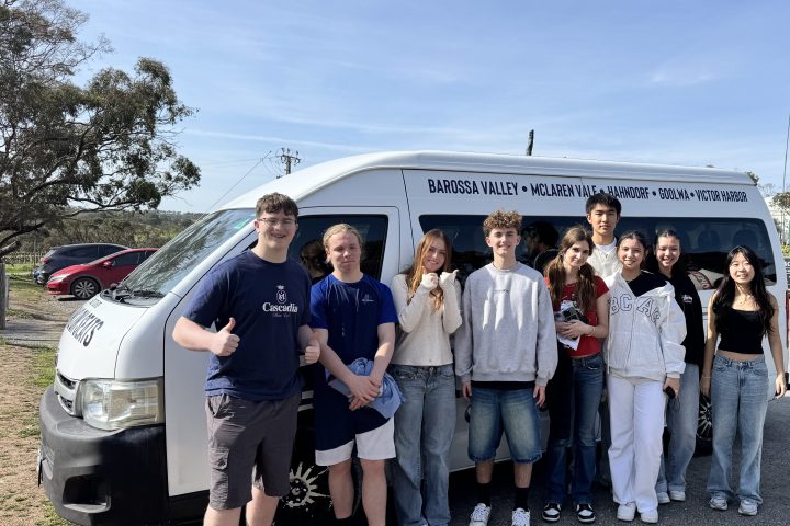 Group of eight friends standing in front of a tour van and sculpture, outdoor setting.