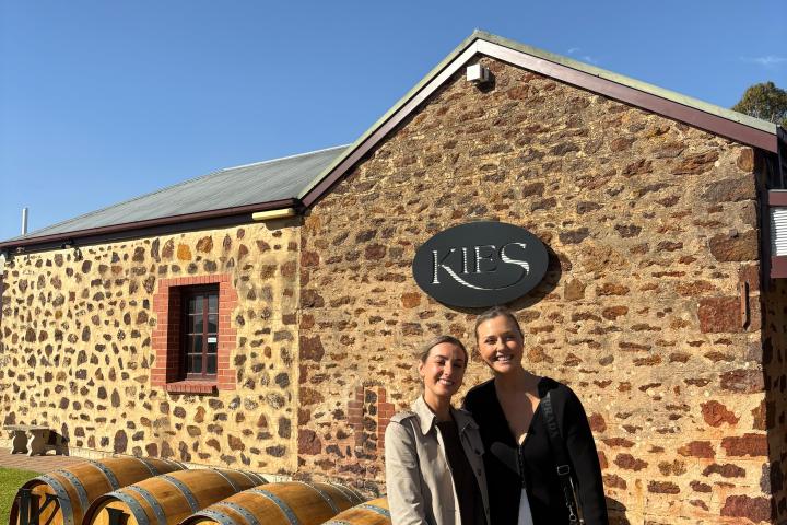 Two people standing by a winery building with wooden barrels and the word 'KIES' visible.