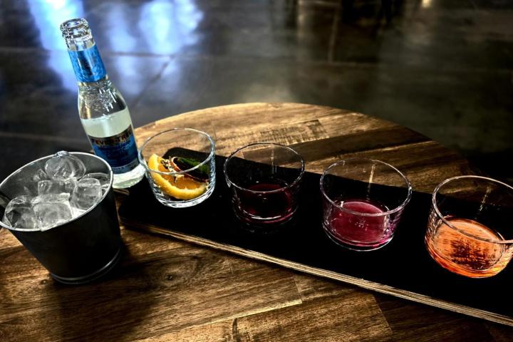 Tray with four glasses of colored drinks, bottle, and ice bucket on wooden table.