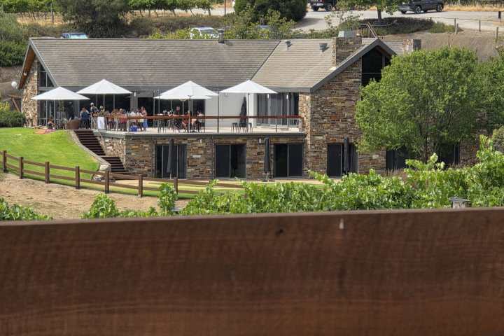 Stone building with patio umbrellas and people, surrounded by vineyards.