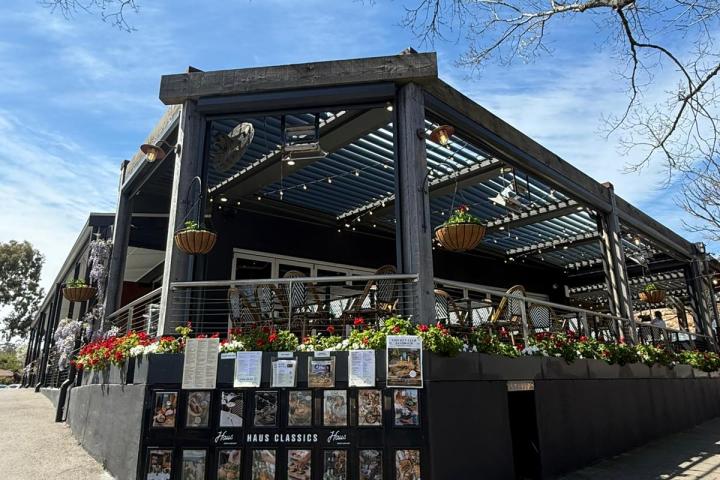 Outdoor cafe with wooden pergola, hanging plants, and menu posters on black walls under blue sky.