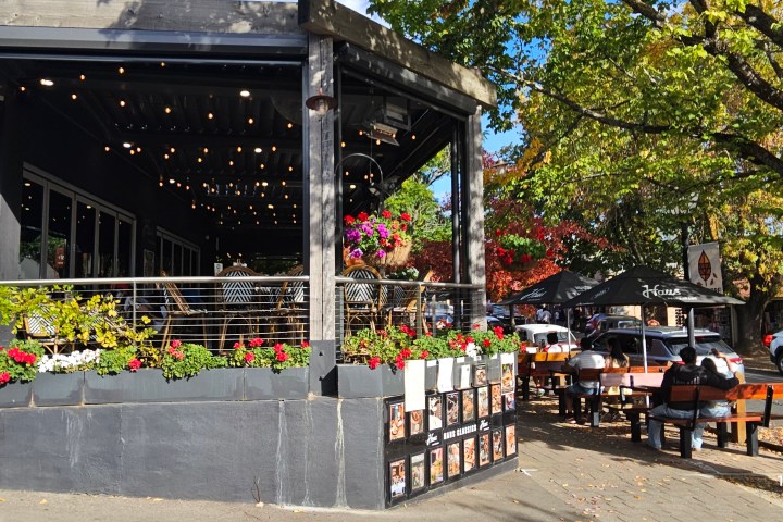 Outdoor café with string lights and flower baskets, people eating at wooden tables under trees.