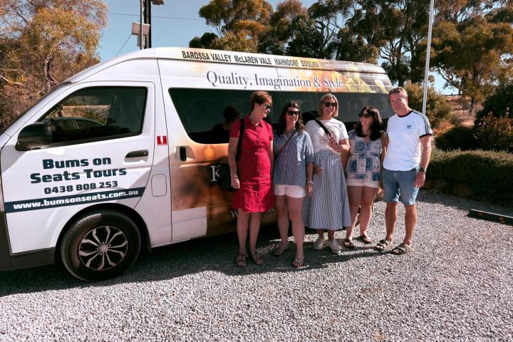 Five people standing beside a tour van labeled 'Bums on Seats Tours' on a gravel lot.