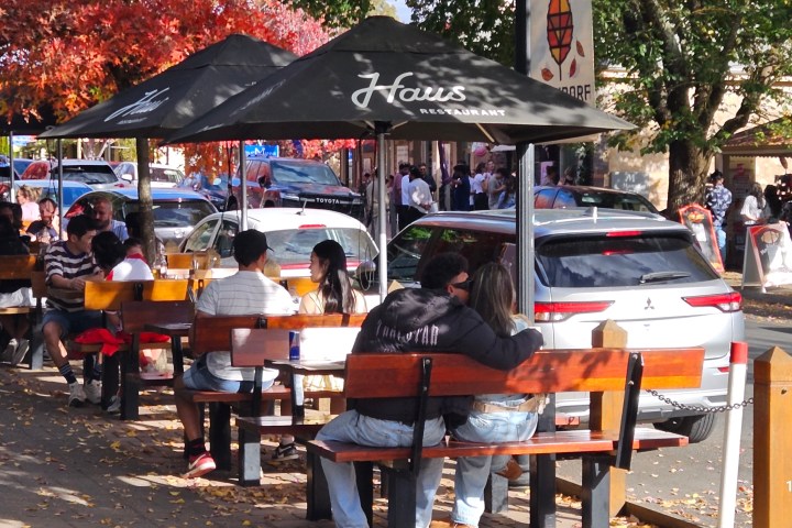 People dining outdoors under umbrellas with autumn leaves around, cars parked nearby on the street.