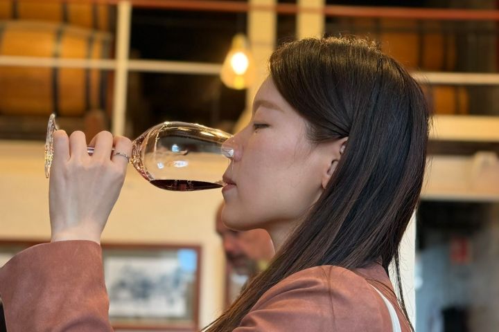 Woman in a pink jacket tasting red wine in a dimly lit room with wine barrels.