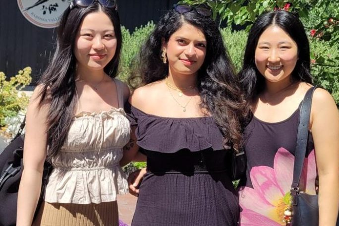 Three women posing outdoors on a sunny day with greenery and flowers.