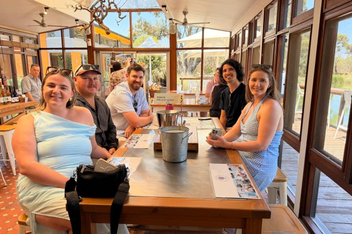 Group of people sitting at a long table with large windows and greenery outside.