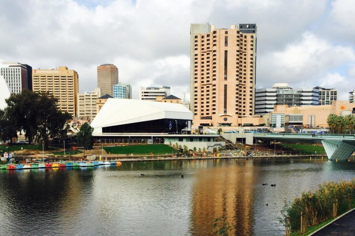 City skyline with tall buildings and a river in the foreground under a cloudy sky.