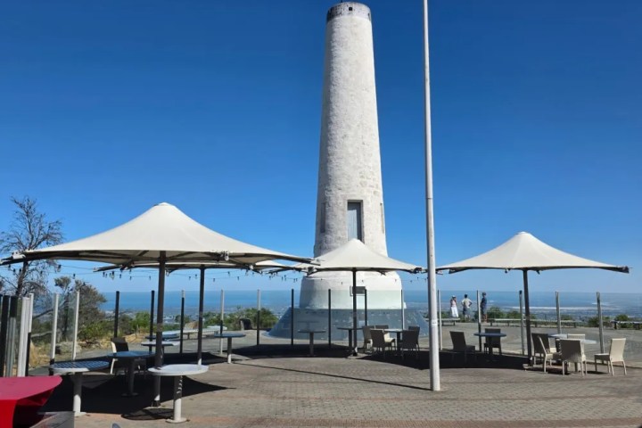 Tall white tower near outdoor seating and umbrellas under a clear blue sky.