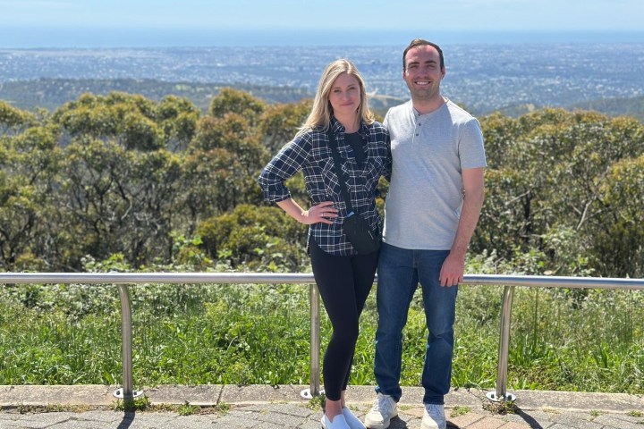 Two people posing in front of a scenic overlook with trees and a cityscape in the background.